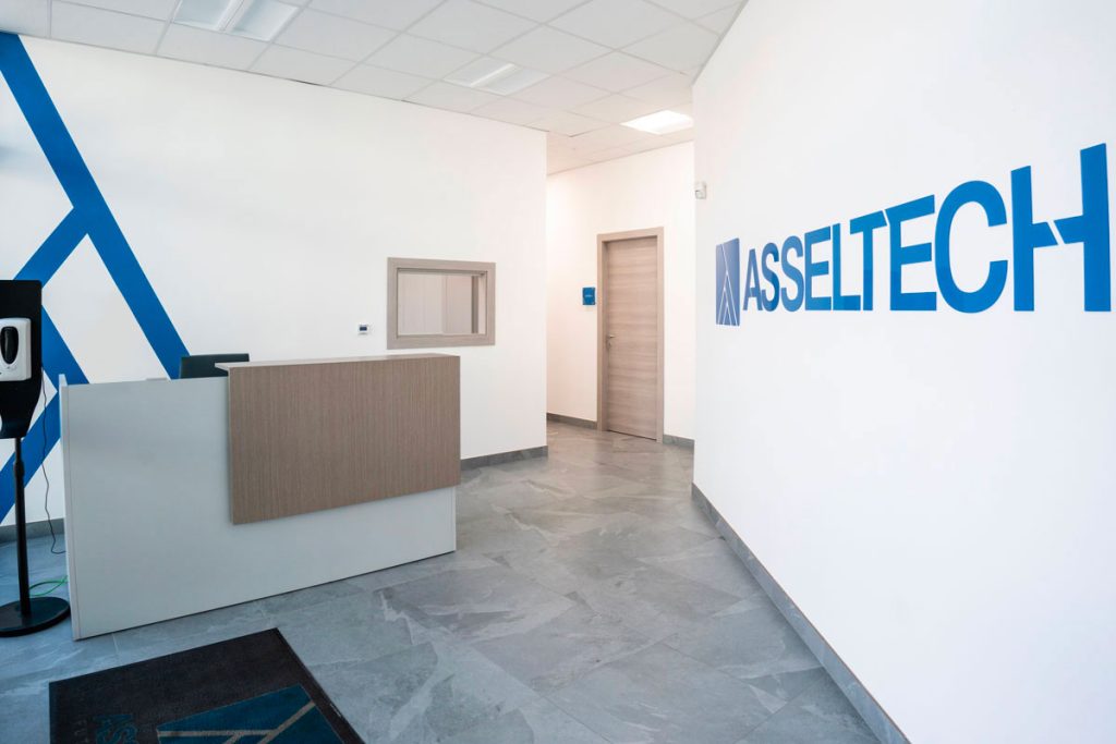 Vista dell'ingresso di un ufficio moderno, con un banco di reception in legno chiaro e un logo aziendale 'ASSELTECH' ben visibile sulla parete. La stanza è luminosa, con pavimento in piastrelle grigie e una parete decorata con una diagonale di linee blu. Si intravede una finestra e una porta, creando un ambiente professionale e accogliente
