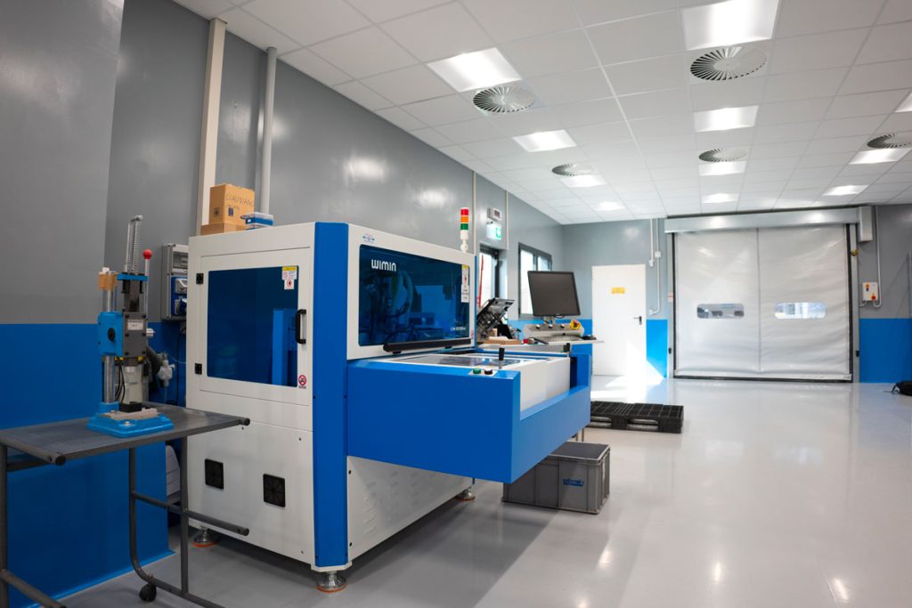 Reparto industriale moderno con una macchina WIMIN per la lavorazione di componenti elettronici. L’ambiente è spazioso, ben illuminato e caratterizzato da superfici pulite e organizzate. Sullo sfondo, una porta industriale avvolgibile e una postazione di lavoro con monitor, a testimonianza di un processo produttivo automatizzato e tecnologicamente avanzato