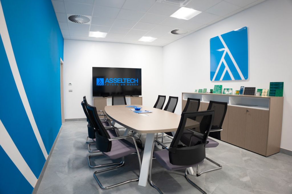 Moderna sala riunioni di Asseltech, caratterizzata da un design minimalista e professionale. Al centro della stanza, un grande tavolo ovale circondato da sedie ergonomiche nere. Sullo sfondo, un monitor con il logo 'Asseltech - Future on Board', mentre sulla parete laterale sono esposti vari circuiti elettronici. L'ambiente è luminoso, con pareti bianche e dettagli in blu, che richiamano l'identità visiva dell'azienda