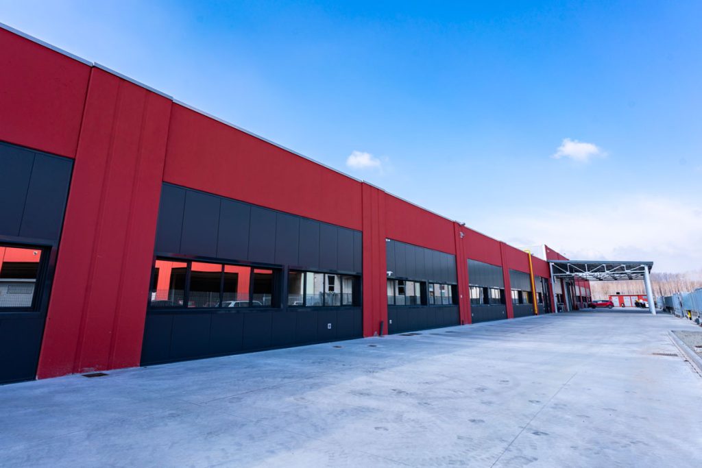 Vista esterna di un moderno stabilimento industriale con pareti rosse e pannelli neri, ampie finestre e una grande area pavimentata. Sullo sfondo, un cielo azzurro con qualche nuvola e una zona di carico coperta con alcuni veicoli parcheggiati