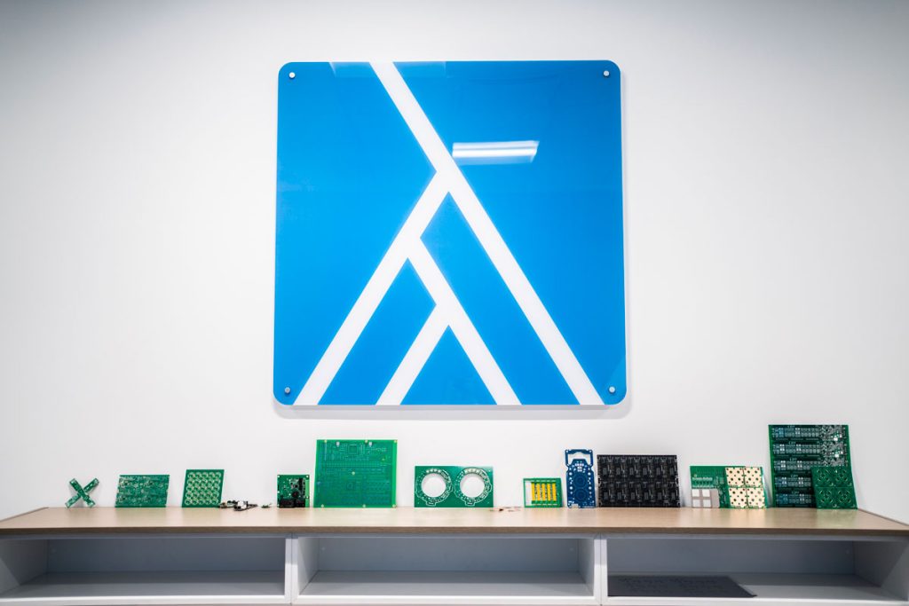 Esposizione di circuiti stampati su una mensola, posizionata sotto un grande pannello decorativo blu con il logo di Asseltech. I circuiti di diverse forme e dimensioni rappresentano l'innovazione e la tecnologia dell'azienda nel settore dell'elettronica. L'ambiente è moderno e minimalista, con un design che enfatizza la precisione e la qualità del lavoro svolto
