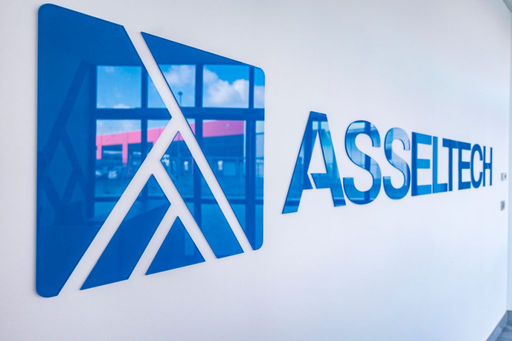 Primo piano del logo Asseltech in rilievo su una parete bianca all'interno di un edificio aziendale. Il logo blu riflette la luce naturale proveniente da una finestra, dove si intravede un'area industriale con strutture e un cielo parzialmente nuvoloso