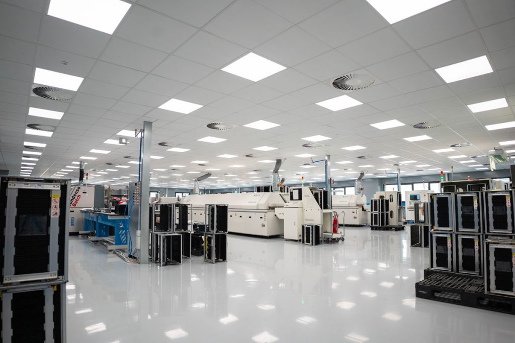 Reparto di produzione high-tech con macchinari automatizzati per l'assemblaggio elettronico. L’ambiente è ampio, luminoso e dotato di sofisticate attrezzature per la produzione di circuiti stampati e componenti elettronici. Il pavimento riflettente e l'illuminazione a LED creano un'atmosfera pulita e altamente tecnologica