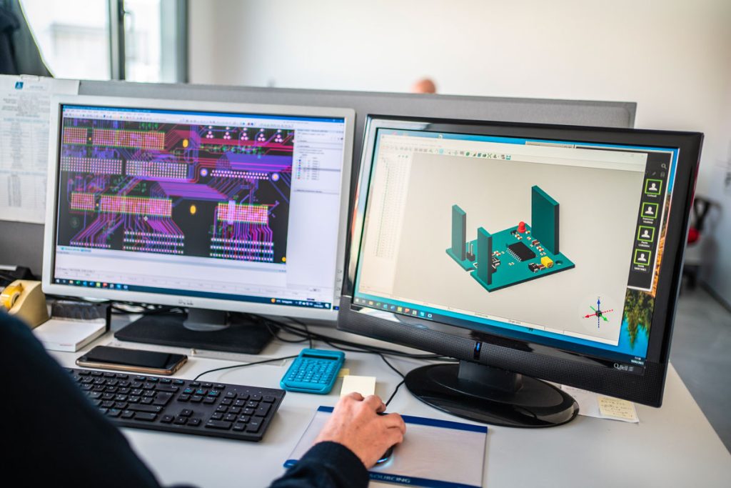 Vista di un workstation con due monitor, uno che mostra un progetto di circuito elettronico su software CAD e l'altro che visualizza un modello 3D di una scheda elettronica. In primo piano, una mano sta usando un mouse su una superficie di appoggio, mentre in fondo si intravede una persona al lavoro