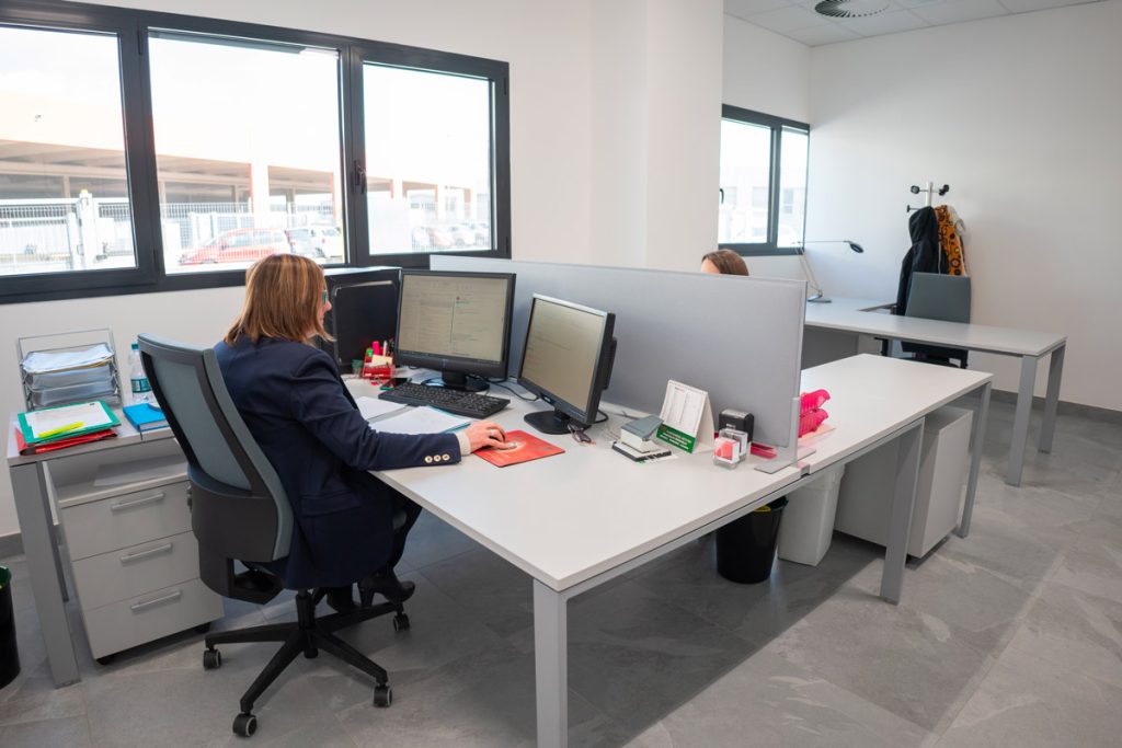 ufficio moderno e luminoso con due scrivanie. Una persona è seduta alla scrivania sulla sinistra, lavorando su un computer. La scrivania è ordinata, con documenti, un bicchiere d'acqua, e materiali organizzati. Si può vedere anche un altro posto di lavoro sullo sfondo, attrezzato con una lampada e una sedia vuota. La stanza ha grandi finestre che permettono l'ingresso di luce naturale