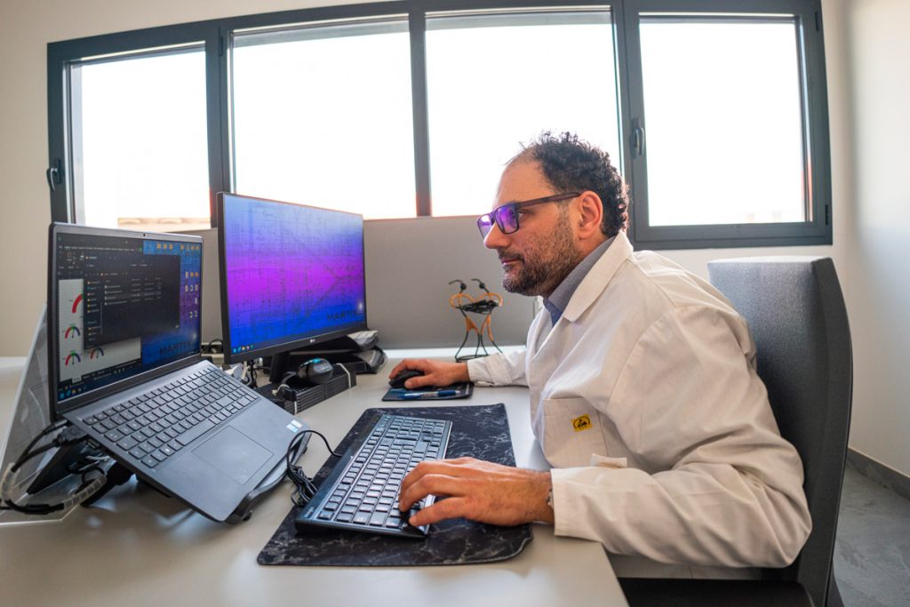 Un uomo in camice bianco lavora al computer in un ufficio moderno, utilizzando due monitor per analizzare dati tecnici. L'ambiente è luminoso, con ampie finestre che offrono luce naturale.