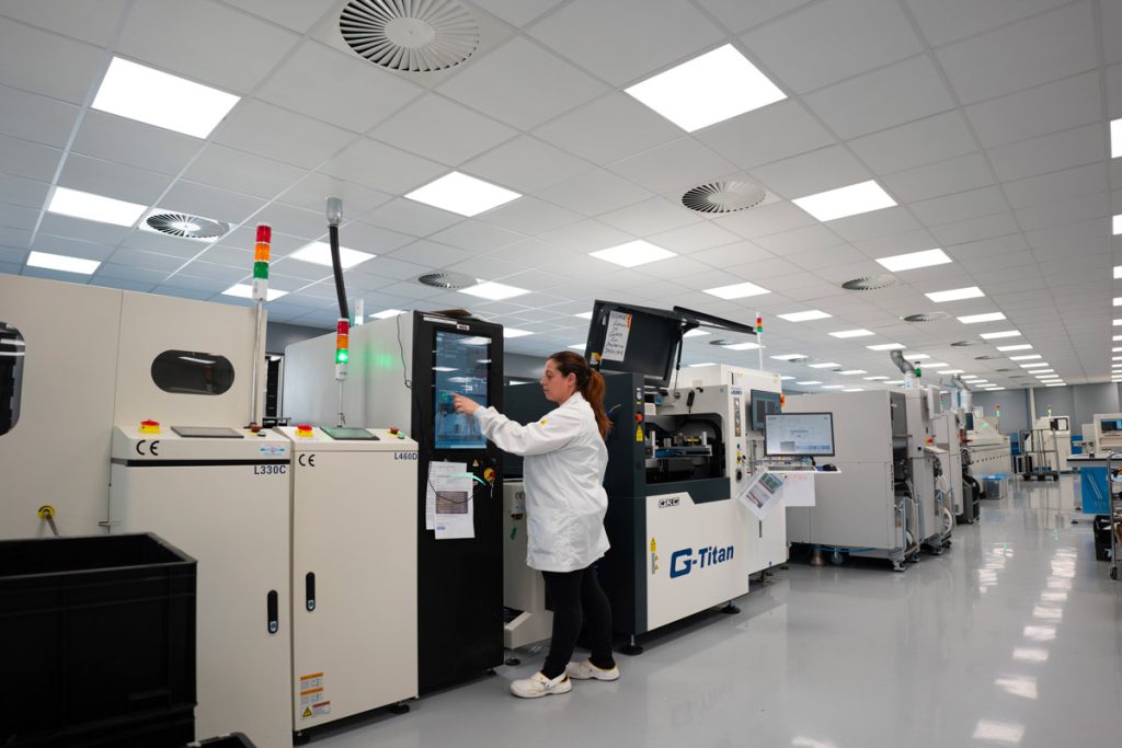 Una donna in camice bianco interagisce con un pannello di controllo su una linea di macchinari industriali in un ambiente di produzione moderno e ben illuminato.