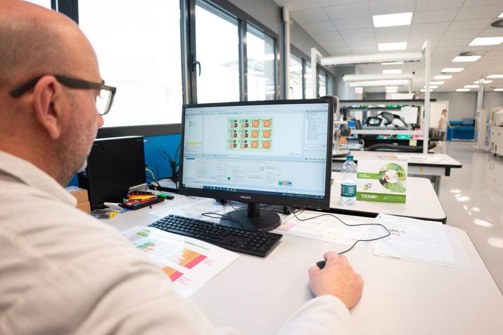 Un uomo lavora al computer in un ufficio moderno, analizzando un progetto su un grande monitor. La scrivania è ordinata, con fogli di dati e un calendario. L'ambiente è luminoso e spazioso, con ampie finestre.