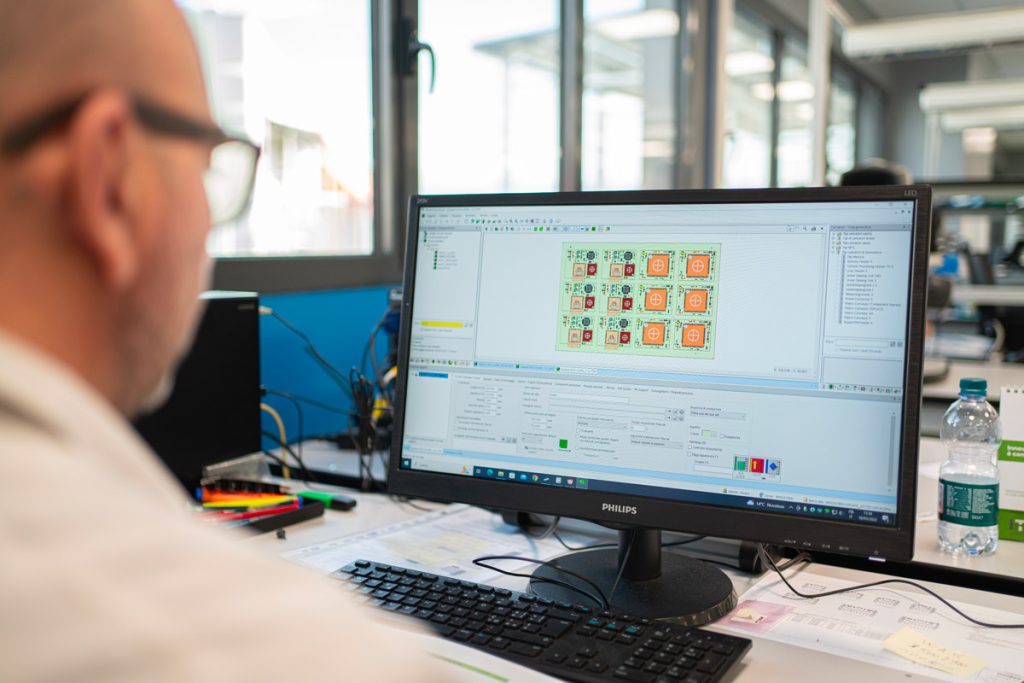 Un uomo lavora al computer, concentrato su un progetto di schede elettroniche visualizzato su un monitor. L'ufficio è moderno e ben illuminato, con finestre che offrono luce naturale.