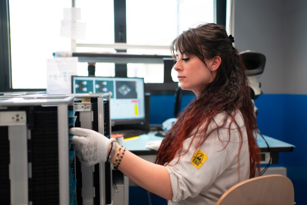 Una donna in camice bianco lavora con attenzione su un dispositivo elettronico in un laboratorio. Indossa guanti protettivi e sullo sfondo si vedono monitor con immagini tecniche. L'ambiente è moderno e ben illuminato.