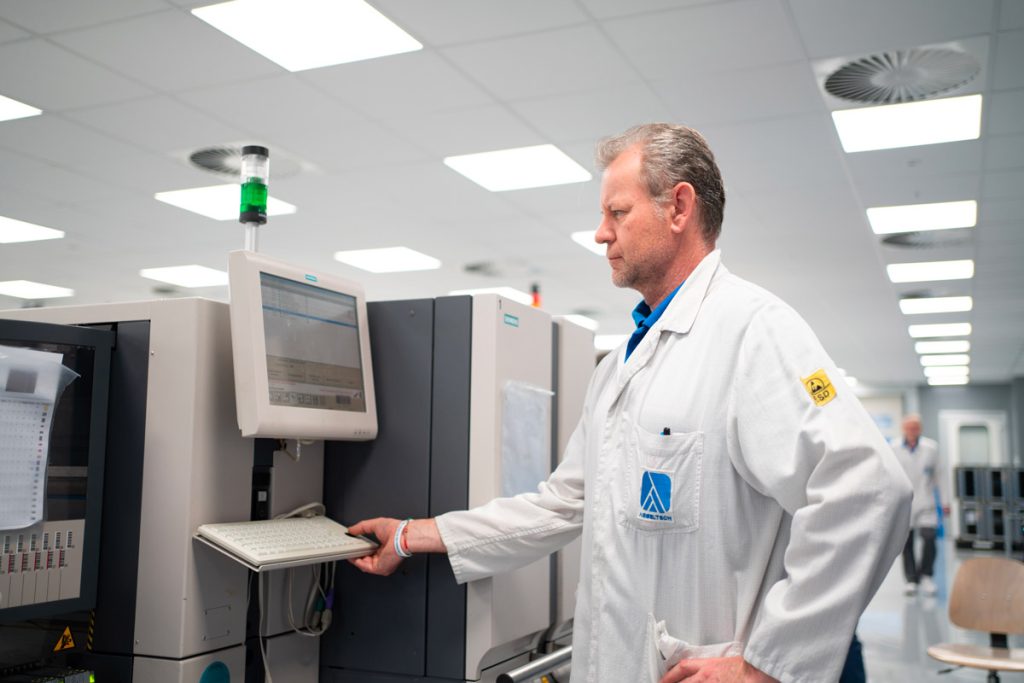 Un uomo in camice bianco interagisce con un monitor e una tastiera su un macchinario industriale in un ambiente di produzione moderno. È concentrato sul suo lavoro e l'area è ben illuminata.
