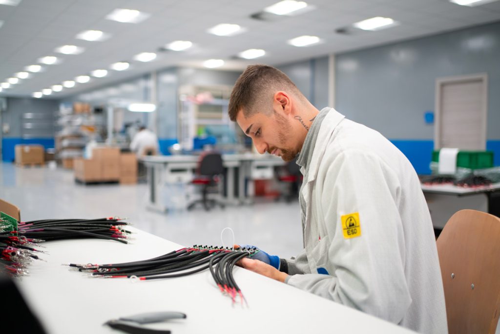 Un tecnico in una fabbrica indossa un camice bianco e lavora concentrato su un gruppo di cavi elettrici su un tavolo. Sullo sfondo si intravedono altri lavoratori e attrezzature industriali.