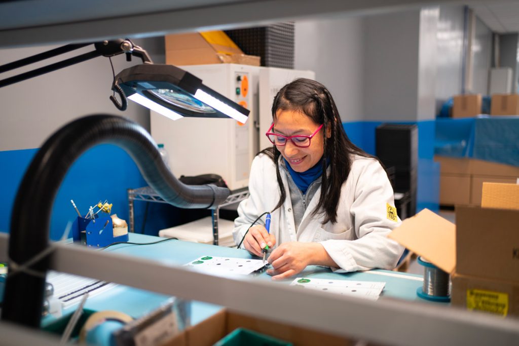 Una donna in laboratorio indossa un camice bianco e occhiali rosa, mentre lavora con attenzione su una scheda elettronica sotto una lampada con lente d'ingrandimento. L'ambiente è ordinato e ben illuminato.