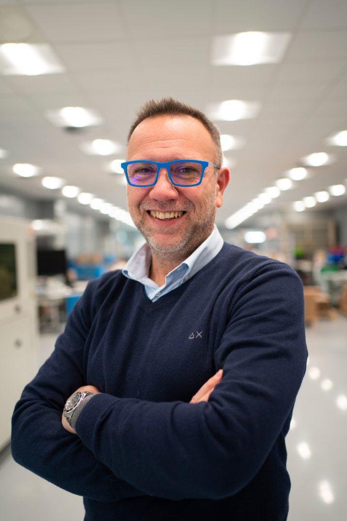 Un uomo sorridente indossa un maglione blu e occhiali azzurri. Si trova in un ambiente lavorativo moderno e ben illuminato, con le braccia incrociate e un'espressione di fiducia.