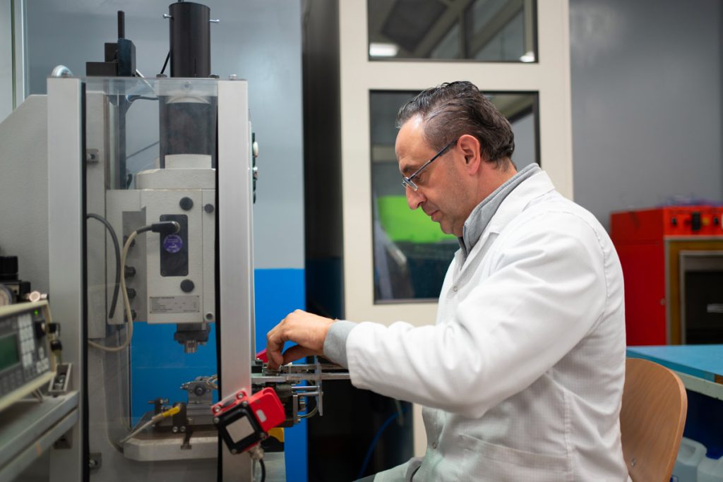 Un uomo in laboratorio indossa un camice bianco e lavora con precisione su un macchinario industriale. L'ambiente è tecnologico e ben organizzato, con apparecchiature moderne.