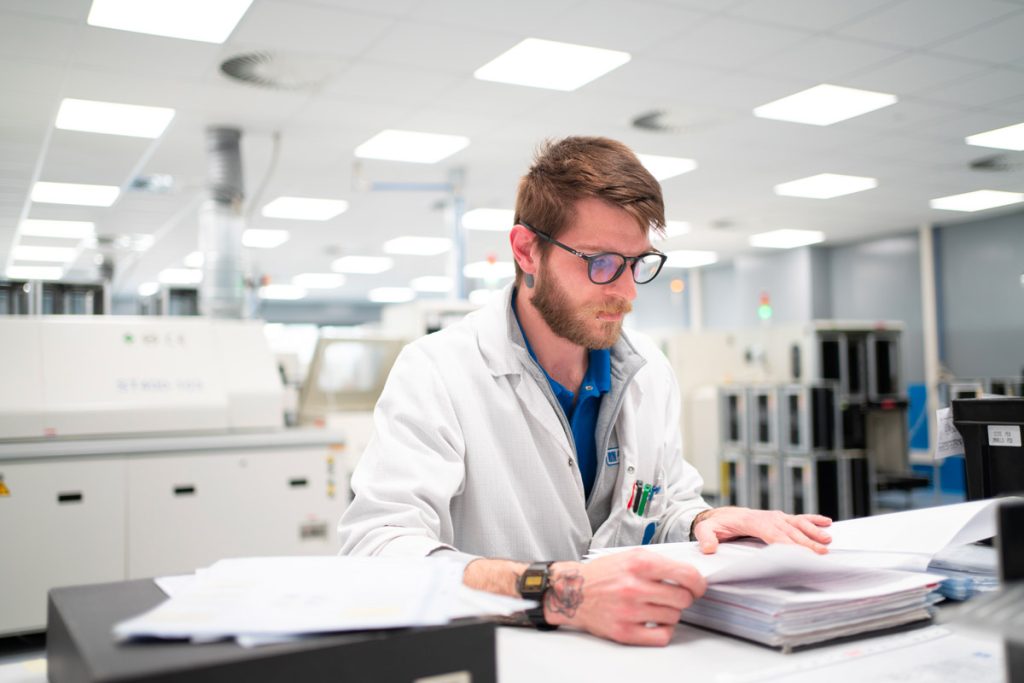 Un uomo con occhiali e camice bianco esamina attentamente documenti su un tavolo in un laboratorio moderno e luminoso. Sullo sfondo si vedono macchinari tecnologici e attrezzature industriali.