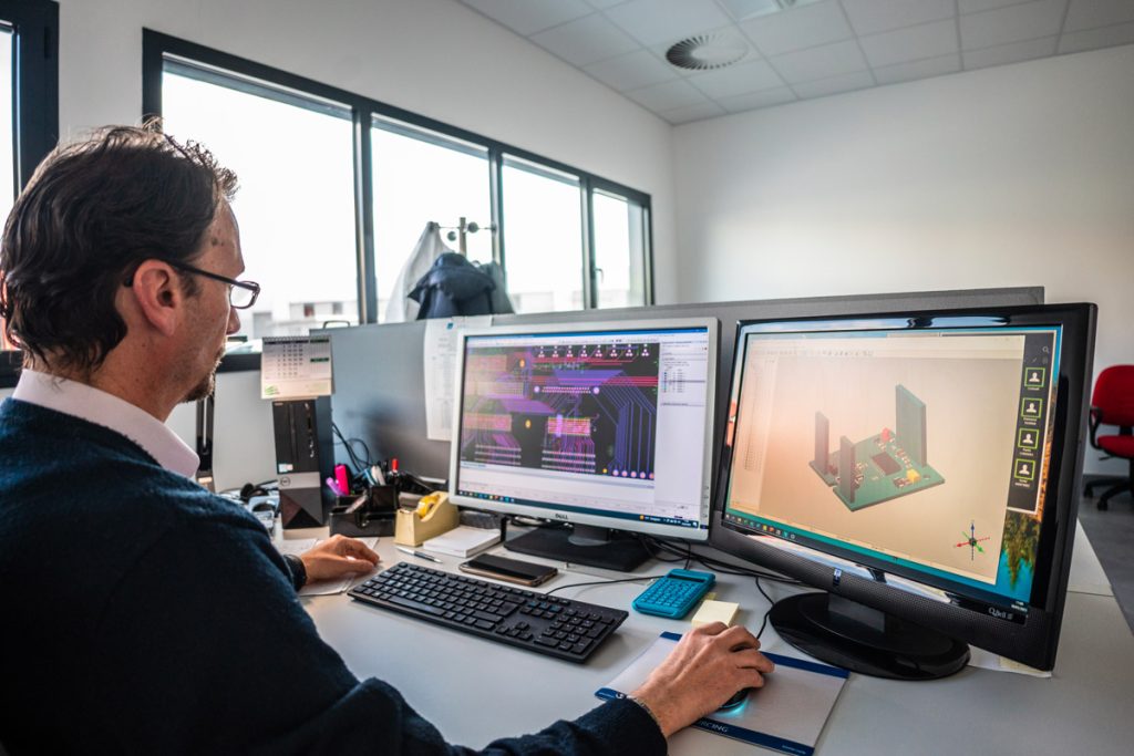 Un uomo lavora al computer in un ufficio moderno, utilizzando due monitor per progettare un circuito elettronico. L'ambiente è luminoso, con grandi finestre che offrono una vista esterna.
