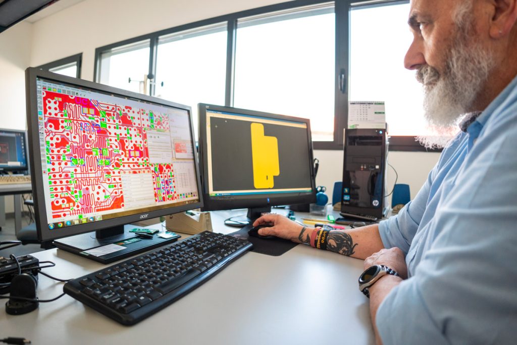 Un uomo lavora al computer in un ufficio, utilizzando due monitor per progettare un circuito elettronico. L'ambiente è luminoso, con ampie finestre che illuminano la postazione di lavoro.